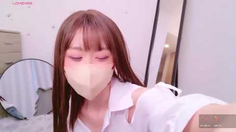 Media: A video of a young Asian woman with straight brown hair and a white surgical mask, wearing a white shirt, sitting on a bed with a mirror and a dresser in the background.