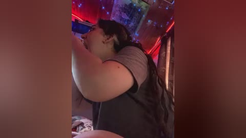 Media: A video of a plus-size person with light skin, wearing a gray shirt, drinking from a bottle in a dimly lit room with red and blue LED lights.