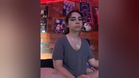 Media: Video of a young woman with light brown skin and long brown hair, wearing a gray shirt, sitting at a table in a dimly lit bar. Brick wall and sports posters in background.