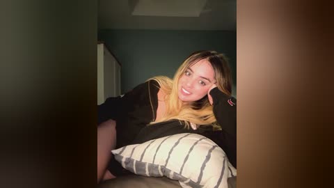 Media: Video of a smiling, light-skinned woman with long blonde hair, wearing a black shirt, leaning on a striped pillow, in a dimly lit room with green walls.