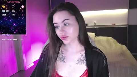 Media: Video of a fair-skinned, long-haired woman with a medium build, wearing a black robe over a red bra, revealing intricate chest tattoos. Background features a modern bedroom with purple and white lighting, and a bed.