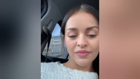 Media: A video of a young woman with fair skin, brown eyes, and straight dark hair, smiling slightly, wearing a white knit sweater. She is seated in a car, with a gray interior and a building visible through the window.