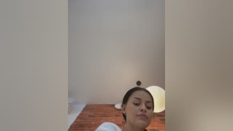 Media: Video of a young woman with a neutral expression, dark hair tied back, standing in a minimalist room with white walls, a red-brick floor, and a large round lamp in the background.