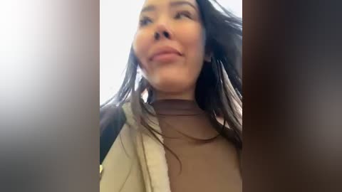 Media: A video of a smiling woman with long, dark hair, wearing a beige jacket over a cream turtleneck, taken from a low angle.