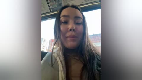 Media: Video of a young Asian woman with long black hair, wearing a beige jacket, smiling inside a bus, with blurred urban scenery visible through the window.