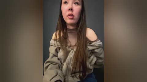 Media: Video of a young woman with long brown hair, wearing a beige off-shoulder top, and blue jeans. She has a pale complexion and a neutral expression. The background is a plain, dark gray wall.