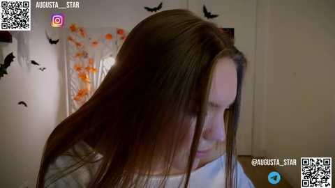 Media: A video of a young woman with long brown hair, wearing a white top, standing in a dimly lit room with Halloween-themed decorations.