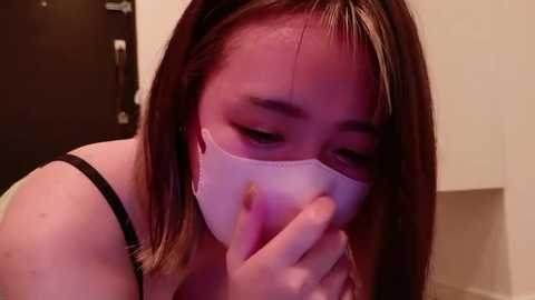 Media: Video of a young woman with light skin and brown hair, wearing a white mask, covering her mouth and nose, and a black bra strap, looking distressed.