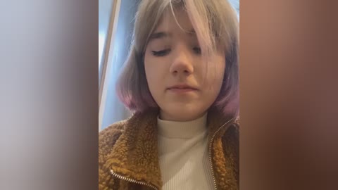 Media: Video of a young Caucasian girl with shoulder-length, pastel pink hair, wearing a brown fuzzy jacket over a white turtleneck. She appears solemn, with neutral expression, in a brightly lit indoor setting.