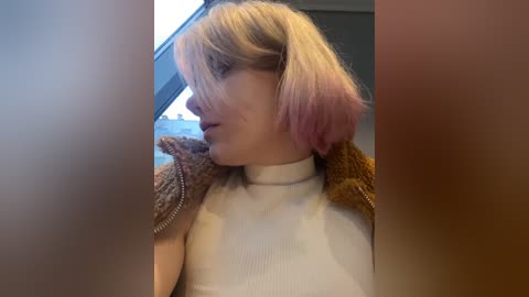 Media: Video of a fair-skinned, blonde woman with a short bob haircut, wearing a white ribbed top and a brown, textured jacket, in a dimly lit room with a window.