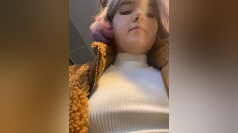 Media: Video of a young woman with pink hair, wearing a white ribbed turtleneck and a brown fuzzy jacket, lying on a bed with a soft teddy bear, in a dimly lit room.