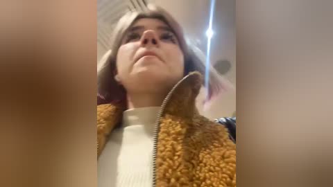 Media: A video of a young woman with light skin and shoulder-length, platinum-blonde hair, wearing a cream-colored sweater and a brown, fuzzy jacket. She gazes upward.