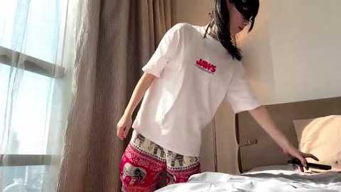 Media: A video of a woman with straight, shoulder-length black hair, wearing a white T-shirt and red and white patterned pajama pants, standing beside a bed in a modern, light-filled bedroom.