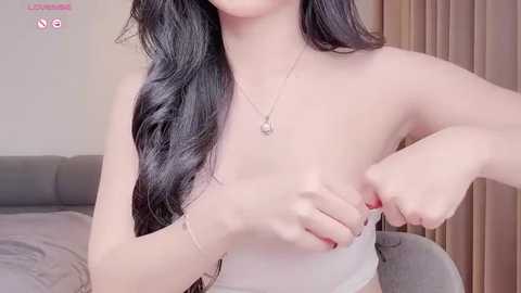 Media: Video of a fair-skinned, slender woman with long black hair, partially undressed, adjusting a white bra, wearing a silver necklace. Background features a grey couch and beige curtains.