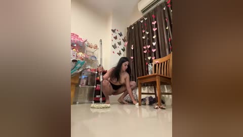 Media: Video of a curvaceous, dark-skinned woman with long black hair squatting on a shiny tiled floor, cleaning with a cloth. Room decorated with heart-shaped decorations, wooden chair, and a whiteboard with colorful scribbles.