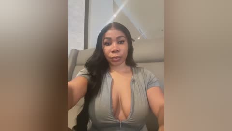 Media: Video of a Latina woman with long black hair, wearing a low-cut grey top that reveals cleavage, sitting indoors on a beige chair.