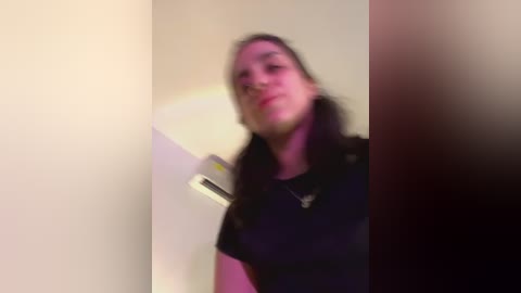 Media: A blurry video of a young woman with long dark hair, wearing a black t-shirt, standing in a dimly lit room with a white wall and a partially visible white object.