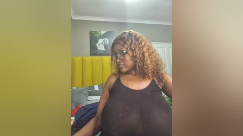 Media: Video of a curvy Black woman with curly brown hair and glasses, wearing a sheer black top, standing in a bedroom with a yellow headboard, a green couch, and a white door in the background.