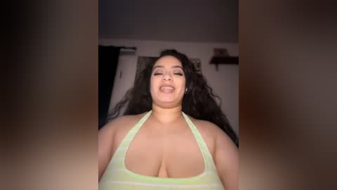 Media: A video of a smiling, curvy Latina woman with long, curly black hair, wearing a revealing neon green halter top, in a dimly lit room with a black curtain and shelves in the background.