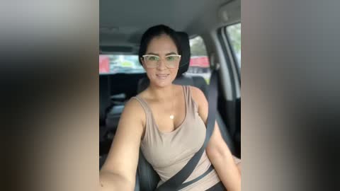 Media: Video of a young woman with medium skin tone, glasses, and black hair, wearing a beige tank top, driving a car.