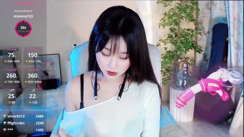 Media: Video of an East Asian woman with long black hair and fair skin, wearing a white off-shoulder top, sitting in a chair, surrounded by plants and a pink teddy bear.