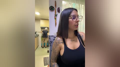 Media: Video of a tattooed woman with long black hair, glasses, wearing a black tank top, standing in a modern kitchen with a man preparing food.