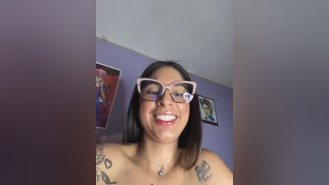 Media: Video of a smiling, topless Latina woman with medium brown skin, wearing glasses, and tattoos on her shoulders. Background includes purple walls with colorful posters.