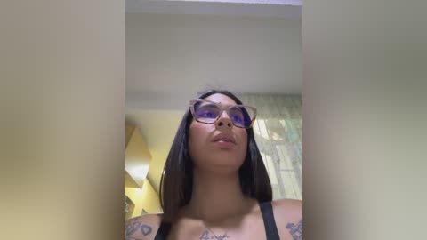 Media: A video of a young woman with medium brown skin, long black hair, and wearing large purple sunglasses. She has tattoos on her arms, wearing a black top, standing indoors with yellow walls and a window with green curtains in the background.