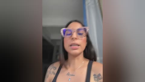 Media: Video of a Latina woman with medium skin tone, dark hair, wearing purple-framed glasses, black tank top, visible tattoos, and a pendant necklace. Background shows blurred indoor setting with white walls.