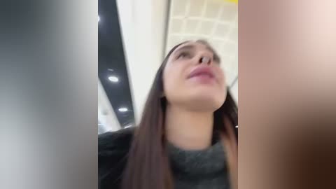 Media: A blurred video of a woman with long brown hair, wearing a black top, looking upward. The background shows a modern, well-lit room with white ceiling tiles. The image is slightly out of focus.