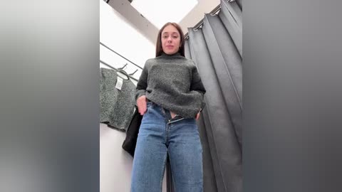 Media: Video of a young woman with light skin and long brown hair, wearing a grey turtleneck sweater and high-waisted blue jeans, standing in a fitting room with grey curtains and a mirror.