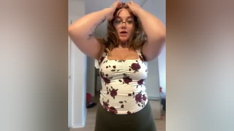 Media: Video of a plus-sized woman with fair skin, glasses, and long wavy brown hair, wearing a white floral top and green pants, standing in a brightly lit room.