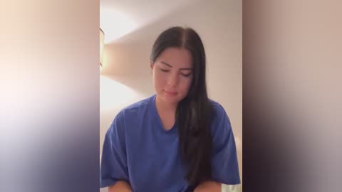 Media: Video of a young woman with long dark hair, wearing a blue hospital gown, standing in a softly lit room with a beige wall. She has a thoughtful expression, looking down.