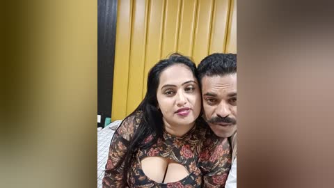 Media: Video of a smiling South Asian woman with medium skin tone, long black hair, and large breasts in a floral top, and a middle-aged man with a mustache, in a yellow room.