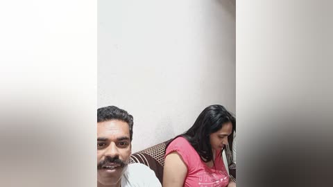 Media: A video showing a smiling man with short dark hair and a mustache, seated next to a woman with long black hair, both wearing casual clothes in a beige-walled room.