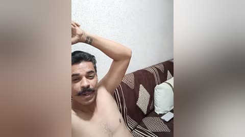 Media: Video of a topless South Asian man with medium skin tone, dark hair, and a mustache, stretching on a bed with patterned brown and white sheets. A white wall and a pillow are visible in the background.