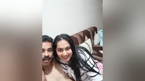 Media: A video of an Indian couple, a man with a mustache and a woman with long black hair, both smiling, on a bed with patterned pillows.