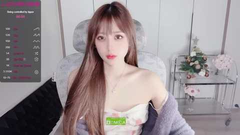 Media: Video of a young Asian woman with long brown hair, wearing a floral off-shoulder top, seated in a modern living room with a glass coffee table and flowers in the background.