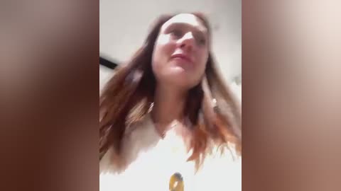 Media: A blurry video of a woman with long, wavy, reddish-brown hair, wearing a white top, captured from a low angle. The background is out of focus, suggesting an indoor setting.