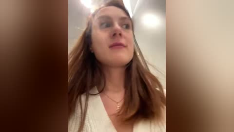 Media: A close-up video of a Caucasian woman with long, straight brown hair, wearing a white top and a gold necklace, looking upward, with blurred faces in the foreground.