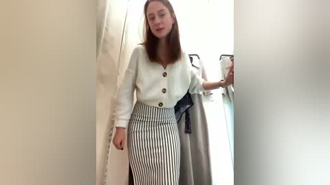 Media: A video of a slim, Caucasian woman with straight, medium-length brown hair, wearing a white, button-down blouse and high-waisted, black-and-white striped pencil skirt, standing in a narrow hallway.