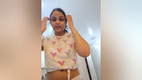 Media: Video of a young woman with medium brown skin and dark hair, wearing a white crop top with pink and purple floral patterns. She is adjusting her glasses in a brightly lit room with white walls.