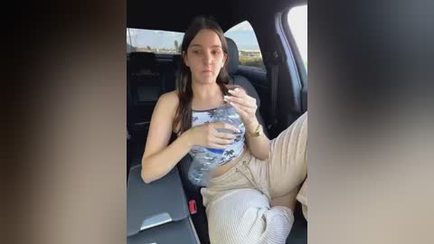 Media: A candid video of a young woman with long dark hair, wearing a camo tank top and beige pants, drinking water from a bottle in the backseat of a car.