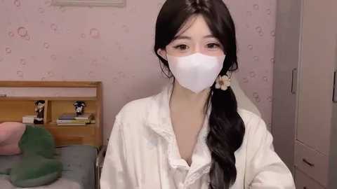 Media: Video of an Asian woman with long black hair in a ponytail, wearing a white mask, white robe, and sitting on a bed with a green pillow. Pink wallpaper with subtle patterns in the background.