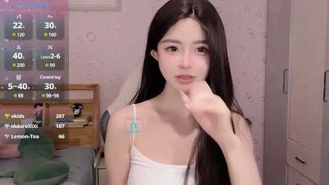 Media: A video of an East Asian woman with long, straight black hair, wearing a white spaghetti-strap top, standing in a pastel bedroom with a bed, wallpaper, and a digital clock overlay.