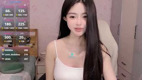 Media: Video of a young, slender East Asian woman with long black hair, wearing a white spaghetti-strap tank top, sitting on a white chair in a bedroom.