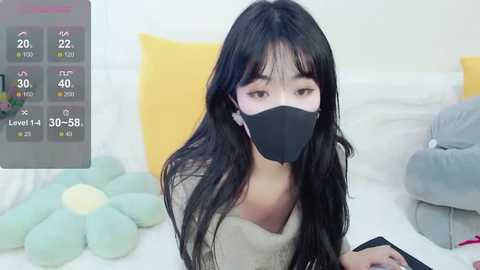 Media: Video of a young Asian woman with long black hair, wearing a black mask and beige sweater, seated on a bed with yellow and green pillows, in a bright room.