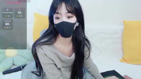 Media: Video of an Asian woman with long black hair, wearing a black mask, gray sweater, and blue jeans, sitting on a bed with yellow and green pillows, in a cozy indoor setting.