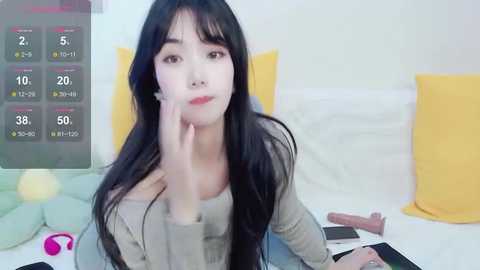 Media: A video of an East Asian woman with long black hair and fair skin, sitting on a bed with a yellow pillow. She has a thoughtful expression, holding a phone, with a digital screen showing game stats in the background.