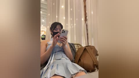 Media: Video of an Asian woman with long black hair, wearing a light blue, sleeveless, pleated dress, taking a selfie with a white iPhone in a room with white curtains and fairy lights.